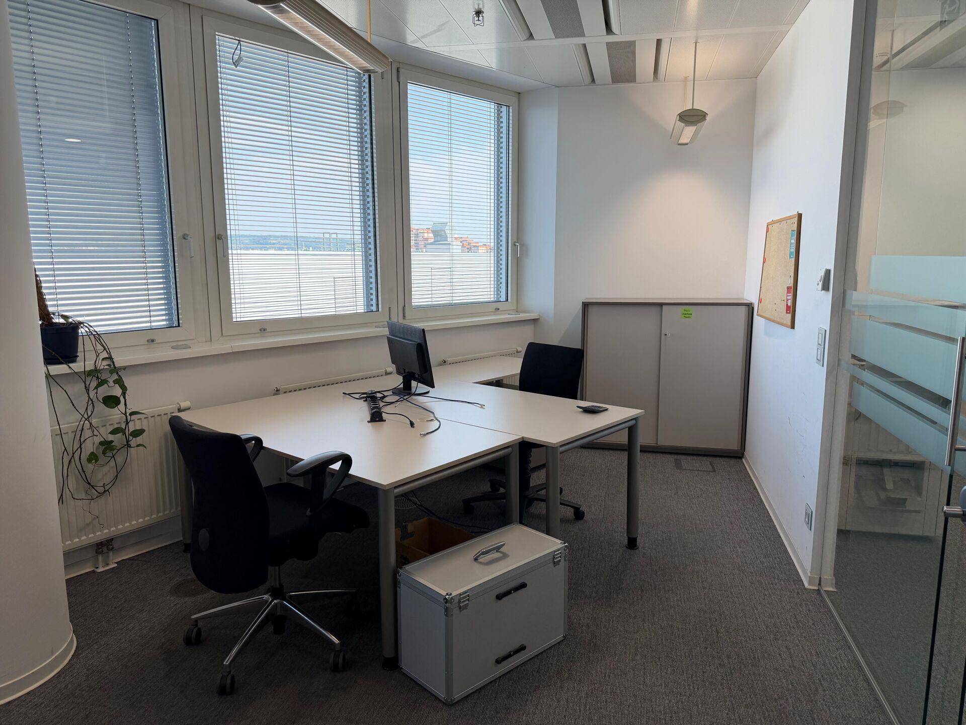 Helles Eckbüro mit zwei Arbeitsplätzen, großen Fenstern und Jalousien.