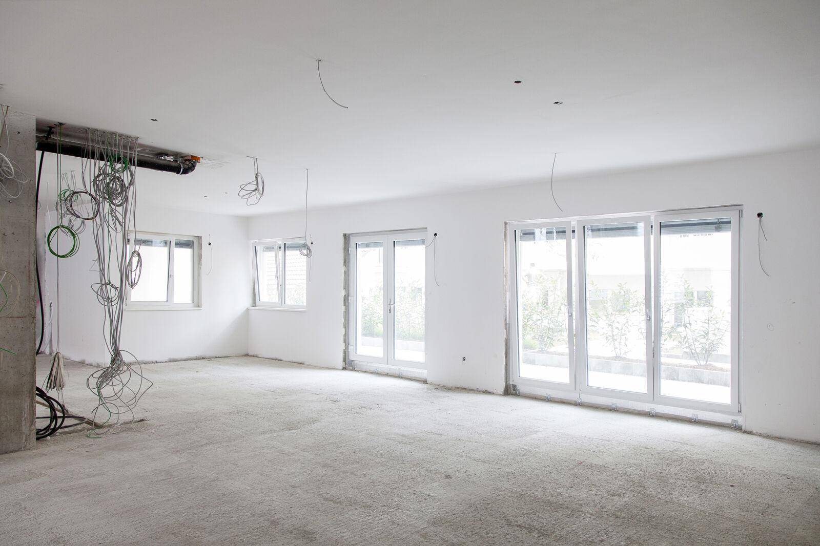 Unfertiger Innenraum im Rohbauzustand mit sichtbaren Kabeln, Betonboden und großen Fenstern für viel Tageslicht.