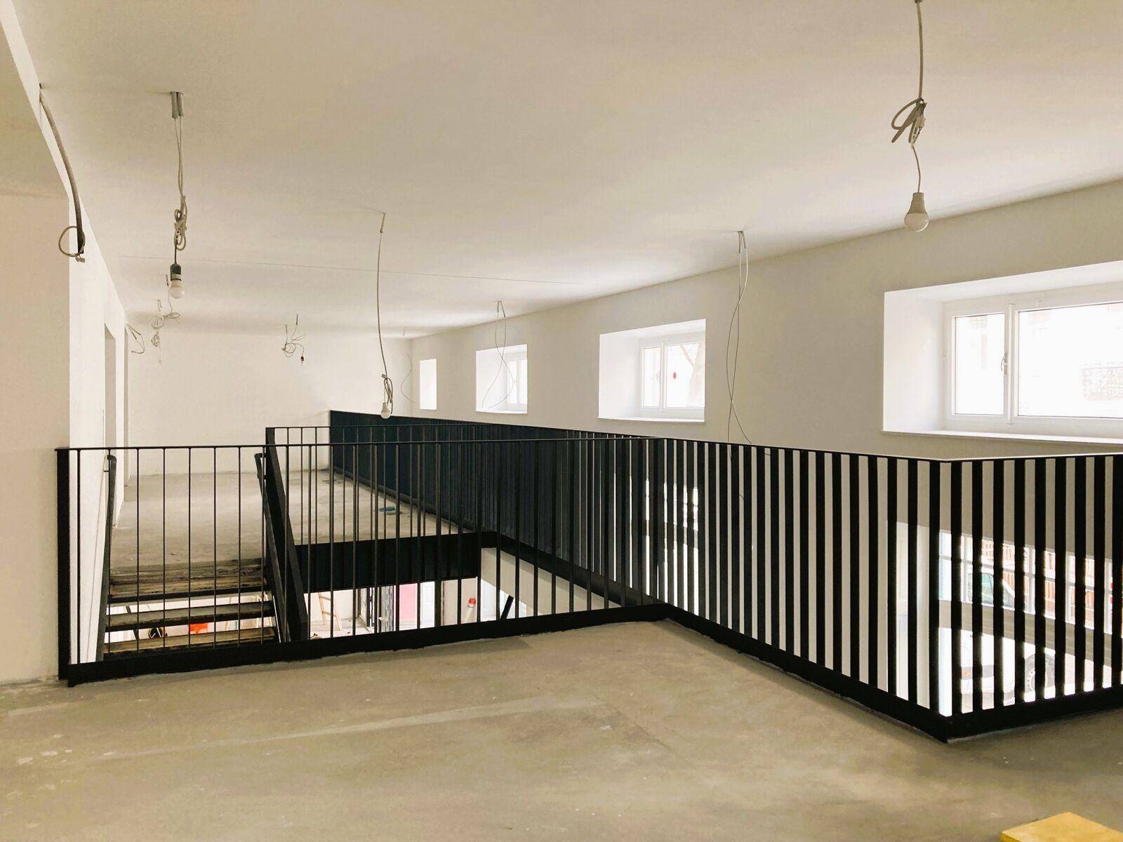 Großer, heller Innenraum im Rohbau mit Galerie, schwarzen Geländern und vielen Fenstern.
