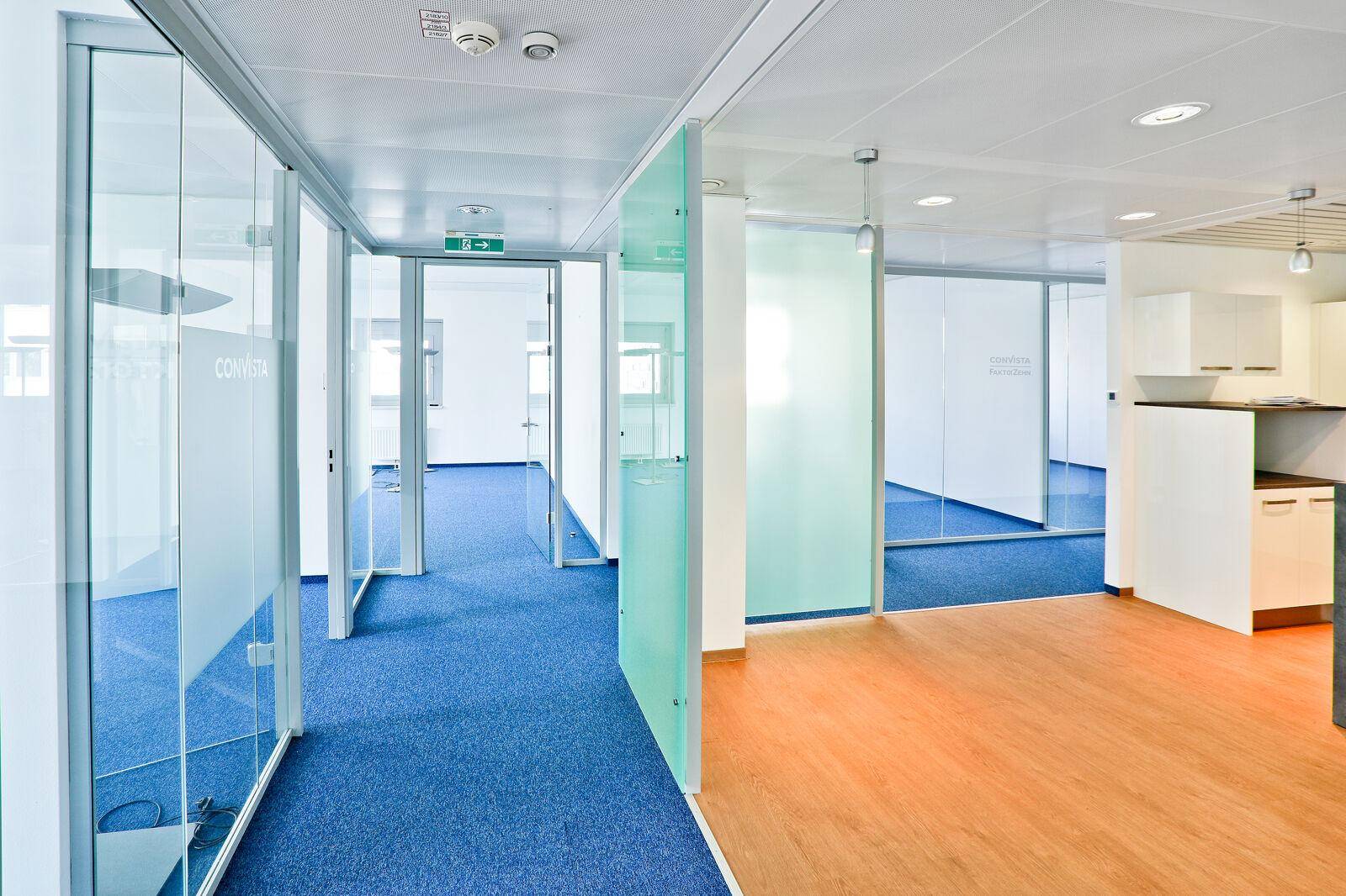 Heller Bürogang mit Glaswänden, der verschiedene Büroräume verbindet und Teppich- sowie Laminatböden aufweist.
