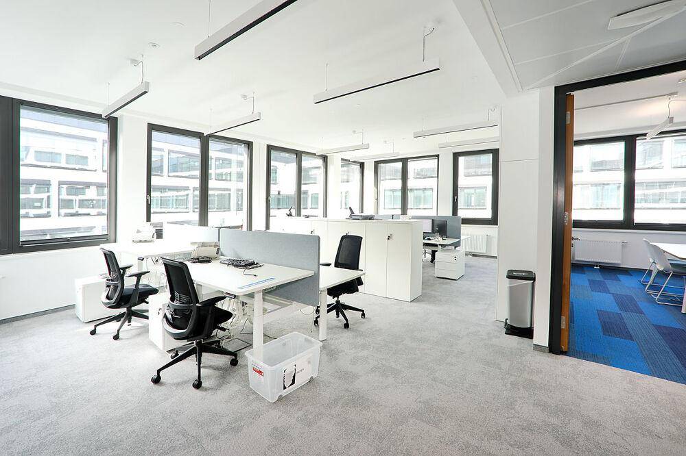 Geräumiges Büro mit mehreren Arbeitsplätzen, großen Fenstern und Teppichboden in hellem Grau.
