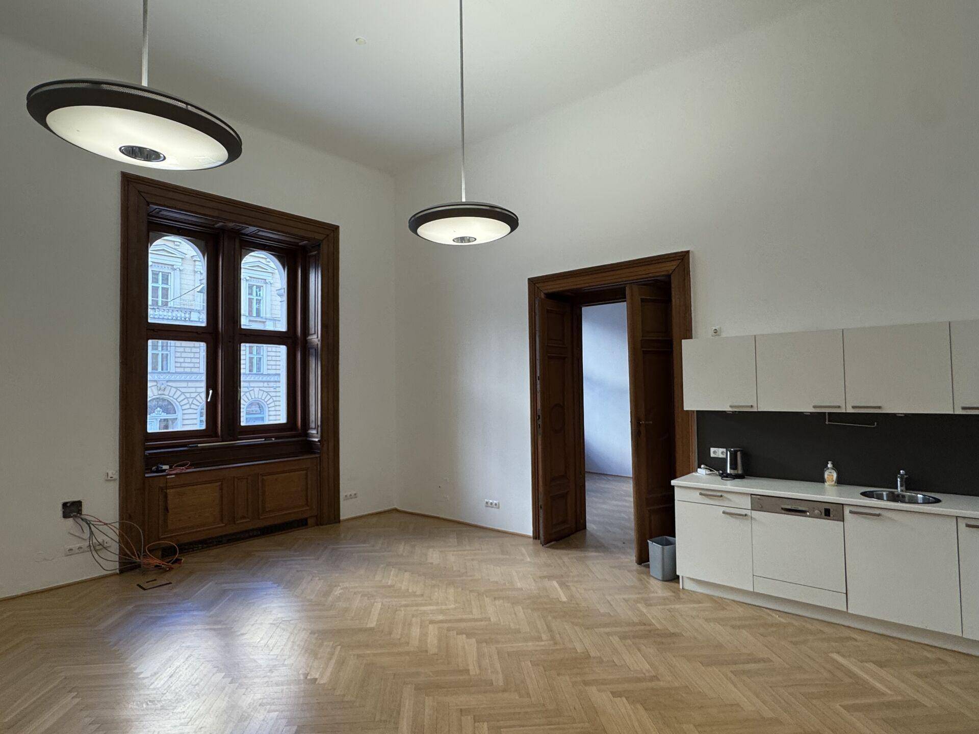 Heller Büroraum mit Fischgrätparkett, hohen Fenstern und einer integrierten, modernen Küchenzeile.