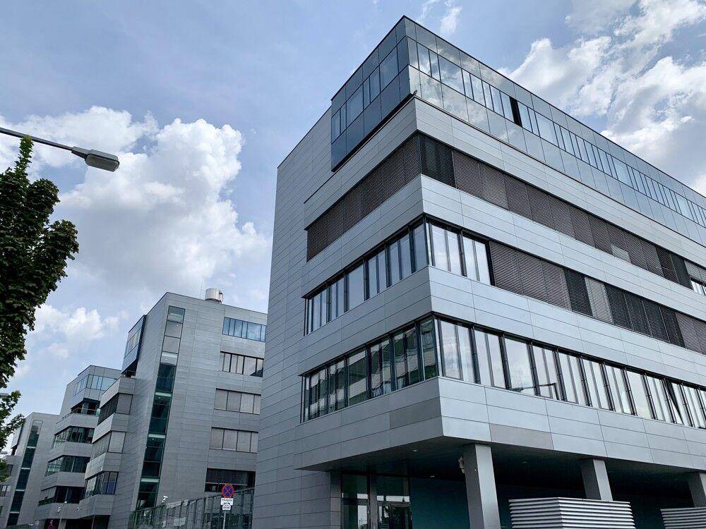 Mehrere moderne Bürogebäude mit Glas- und grauen Fassaden unter einem bewölkten Himmel, mit Bäumen im Vordergrund.