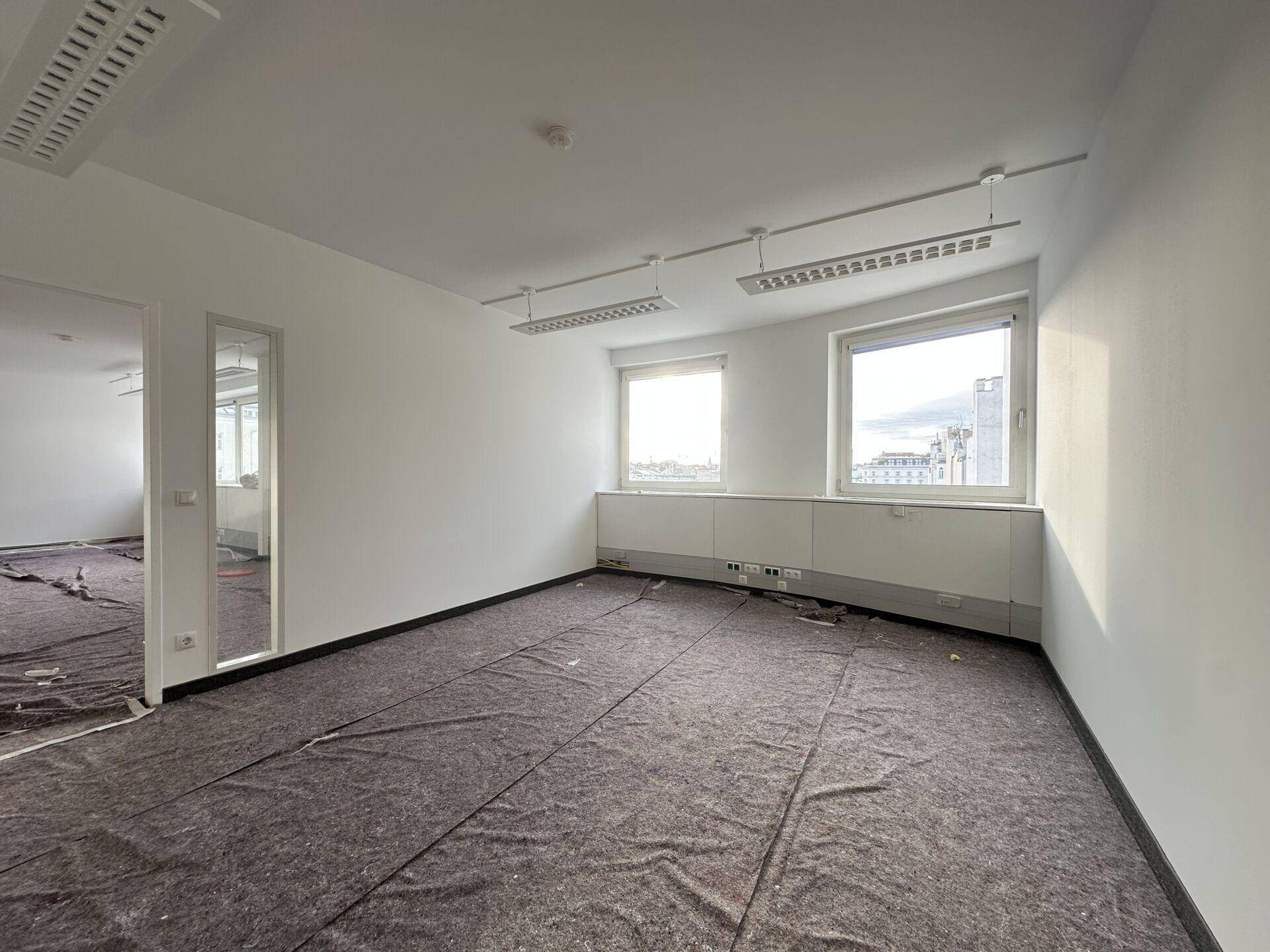 Leerer Büroraum mit schützender Abdeckung auf dem Boden und Fenstern, die einen Blick nach draußen bieten.