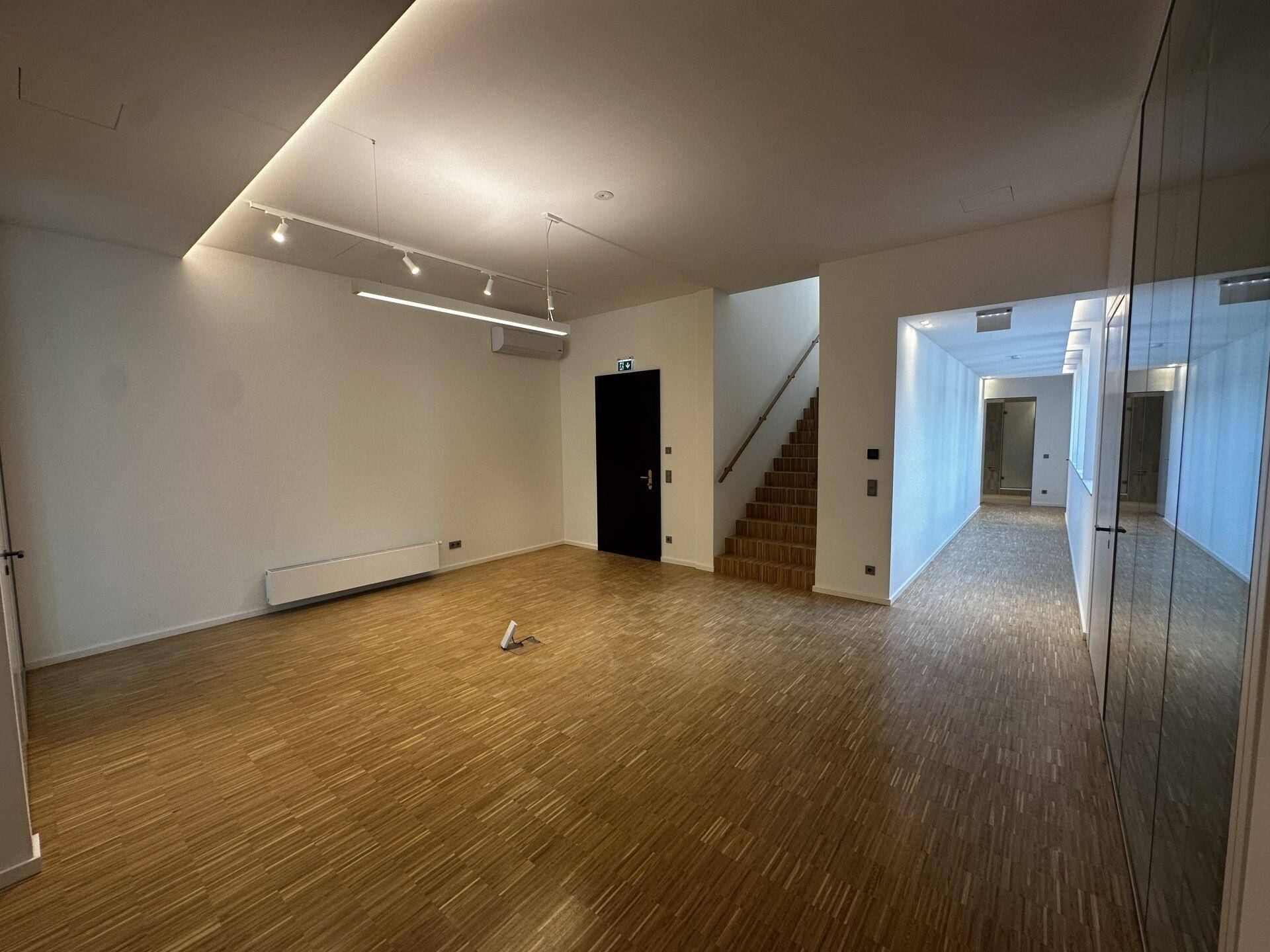 Geräumiger Bürobereich mit Holzparkett, moderner Beleuchtung und Zugang zu einer Treppe und einem Flur.