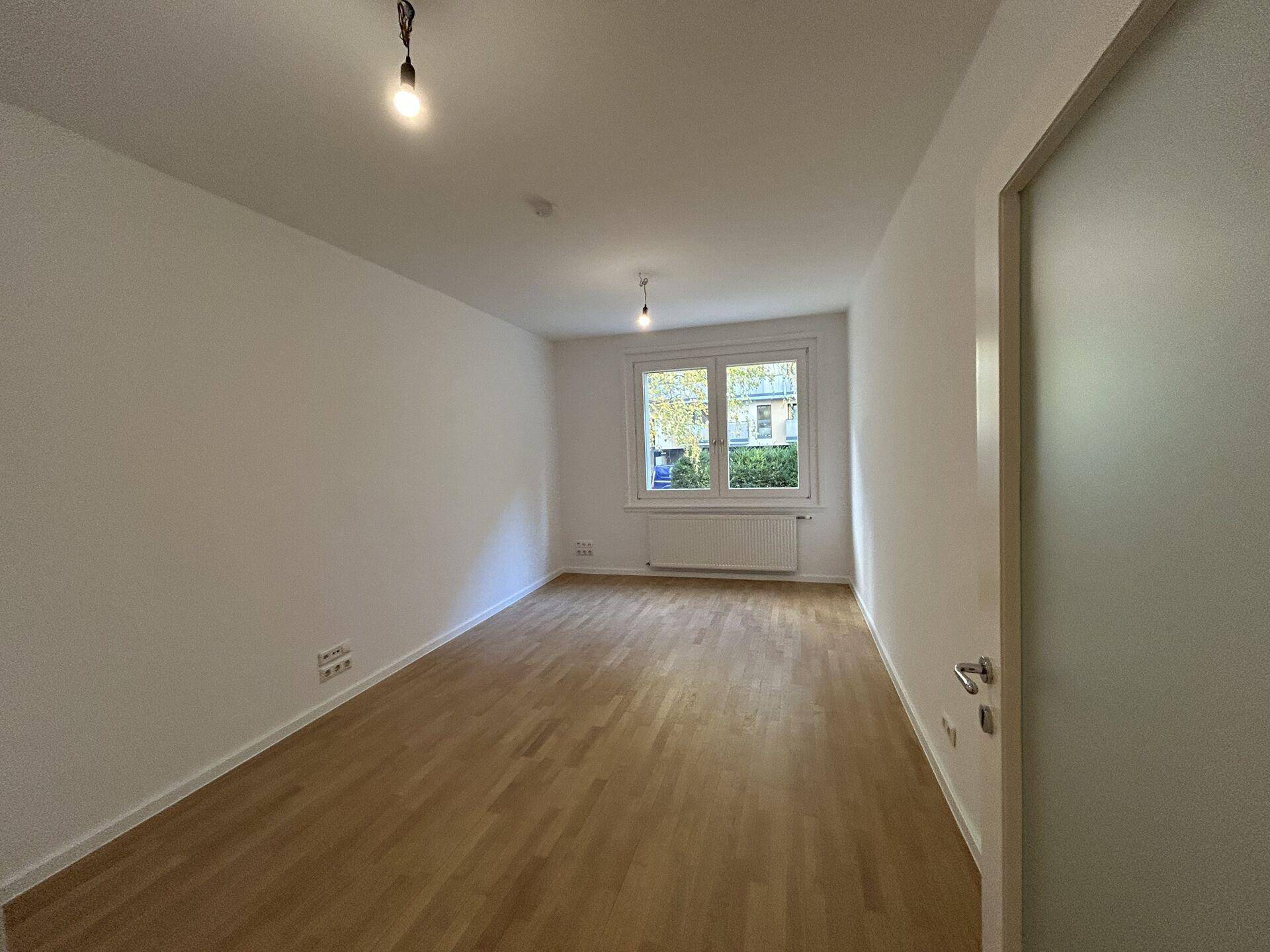 Leeres Zimmer mit hellem Holzboden und weißen Wänden, ausgestattet mit einem Fenster für natürliche Belichtung.
