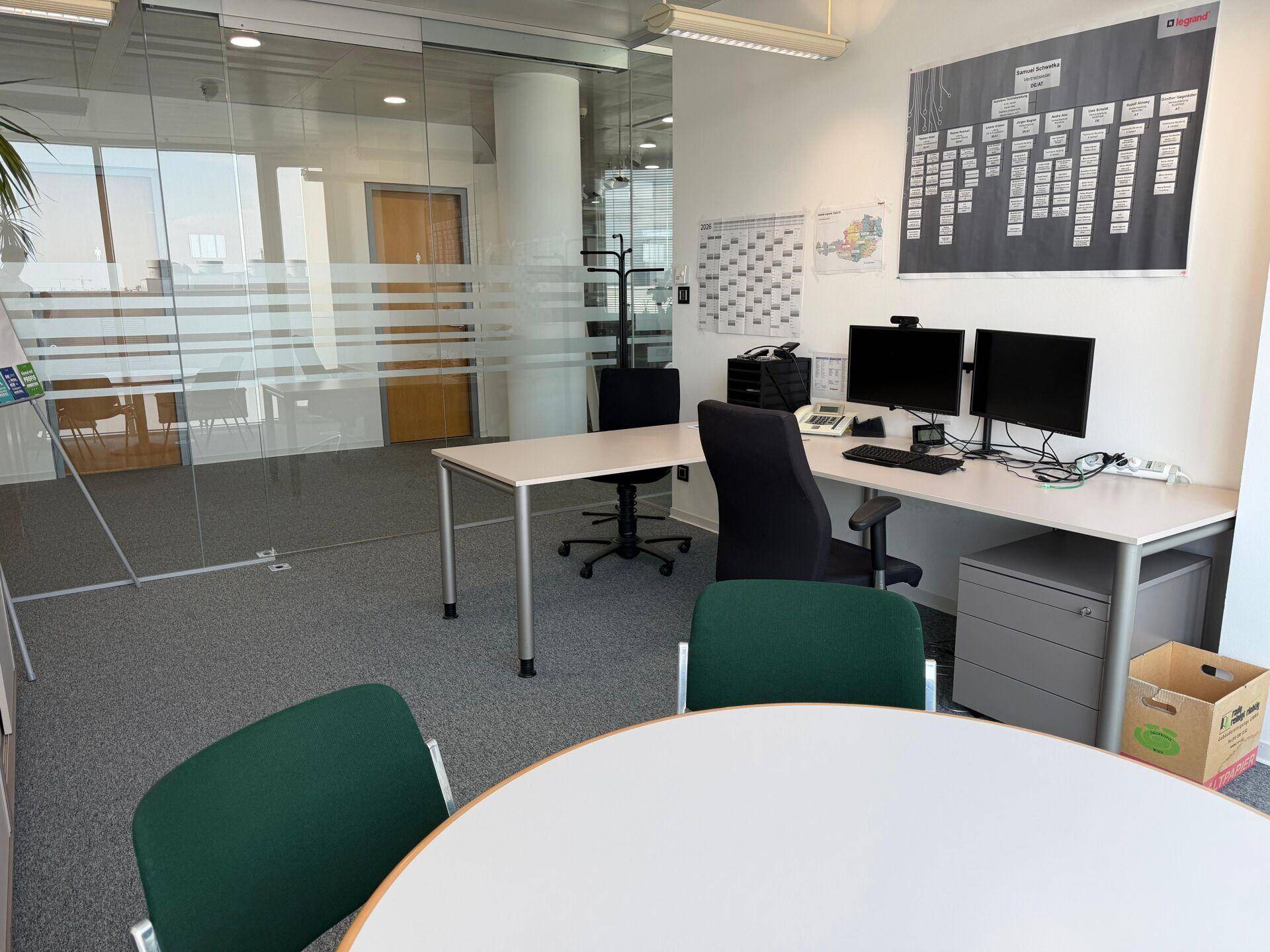 Modernes Büro mit Schreibtisch, Computern und Glaswänden, bietet eine helle und funktionale Arbeitsumgebung.