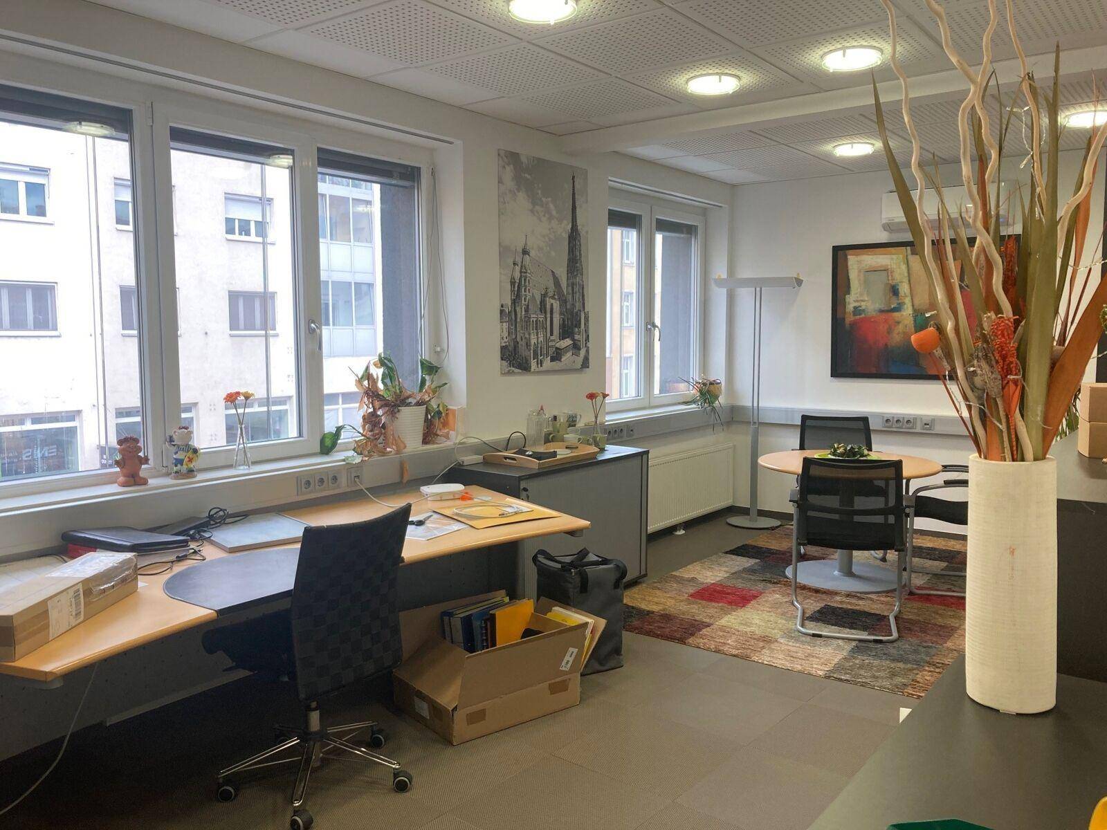 Geräumiges Büro mit mehreren Arbeitsplätzen, großen Fenstern und einem bunten Teppich im Sitzbereich.