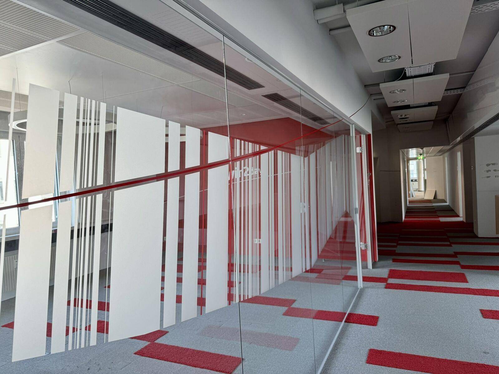 Moderner Bürogang mit Glaswänden, roten Designelementen und Teppichboden.