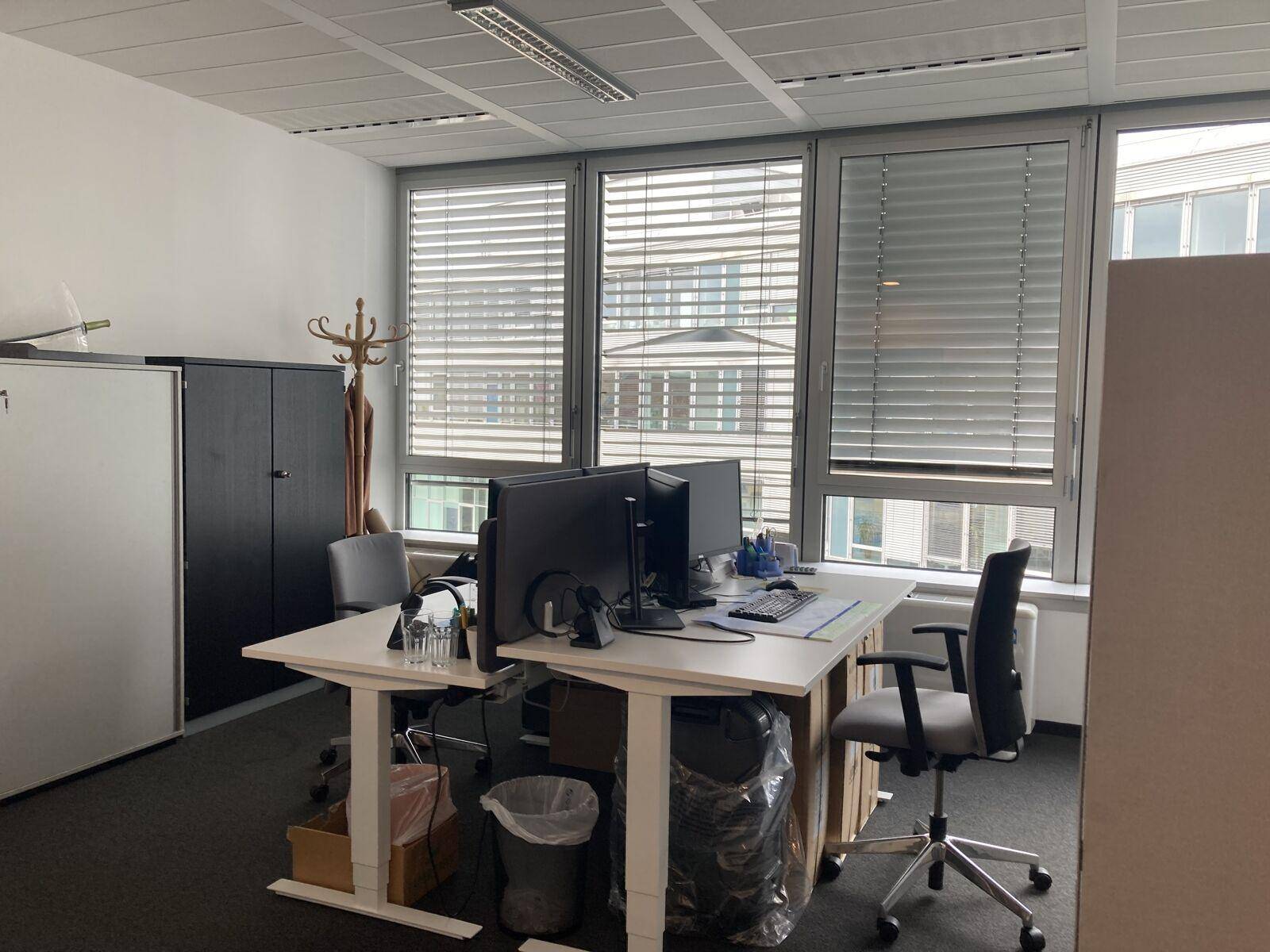 Büroarbeitsplatz mit zwei Schreibtischen, Computern und großen Fenstern mit Jalousien, umgeben von Büromöbeln.