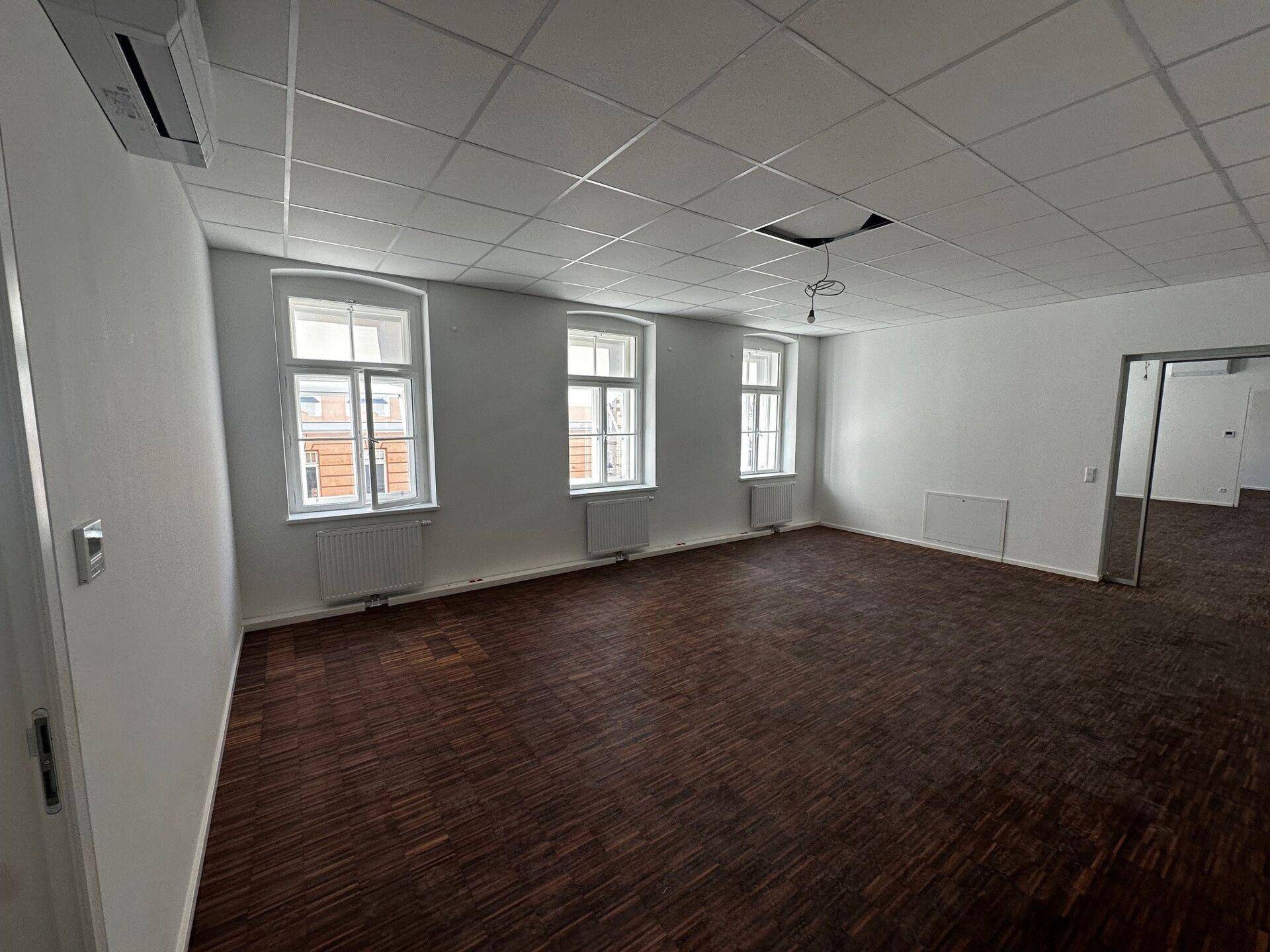 Geräumiges, leeres Büro mit dunklem Holzboden, weißen Wänden und mehreren Fenstern.