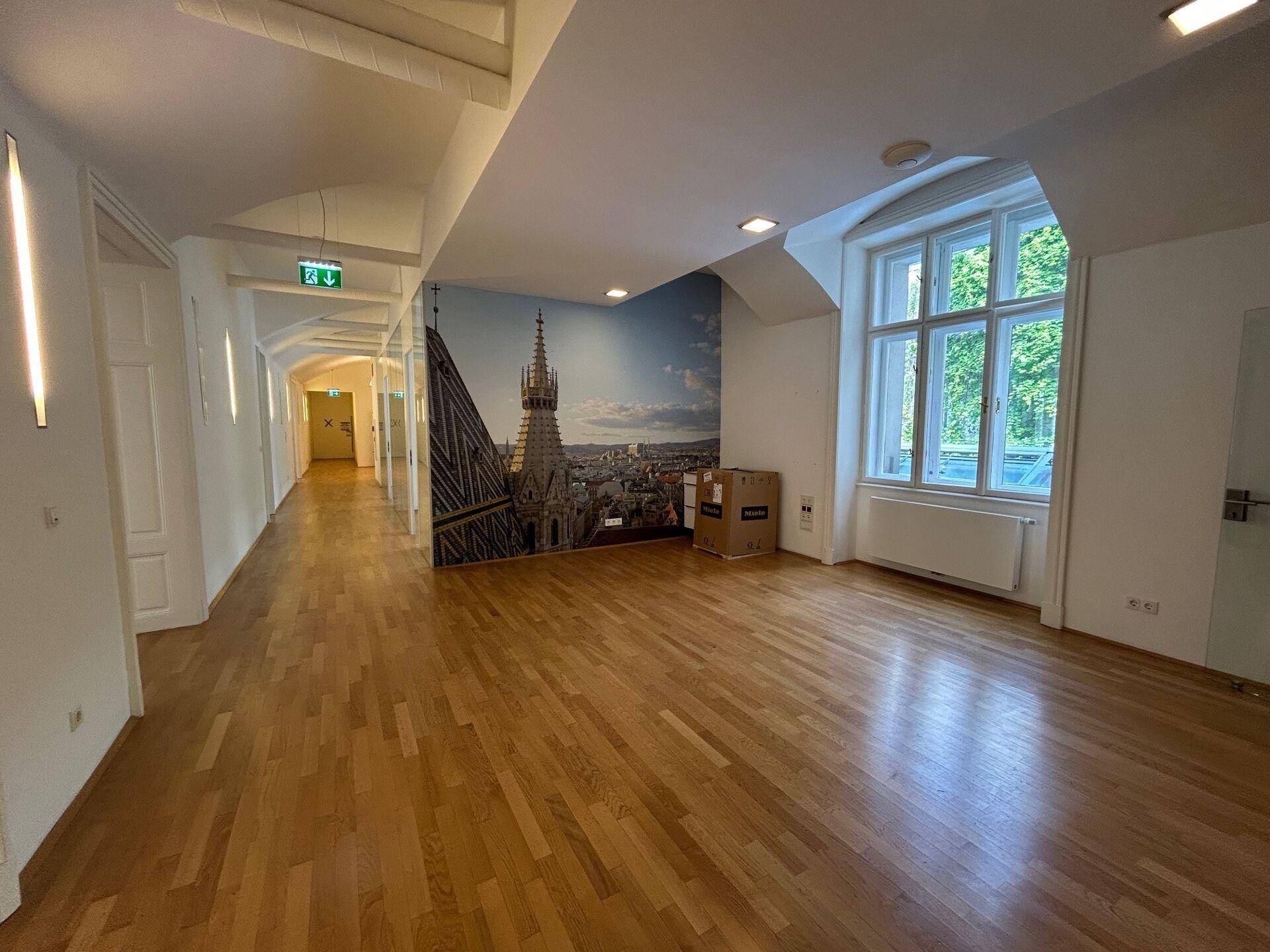 Geräumiger Büroraum mit Fischgrätparkett, großer Fensterfront und einer Wandtapete mit Stadtmotiv.
