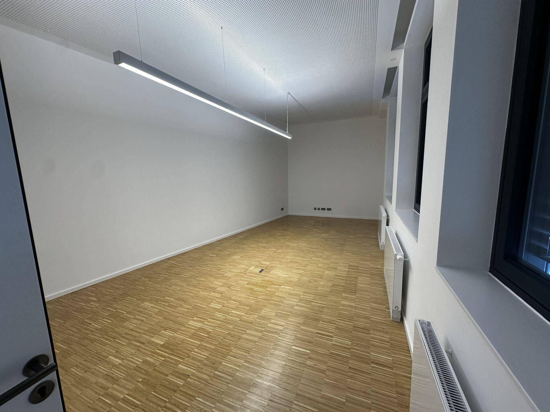 Leerer Büroraum mit Holzparkett, weißen Wänden, Heizkörpern unter den Fenstern und moderner Deckenbeleuchtung.