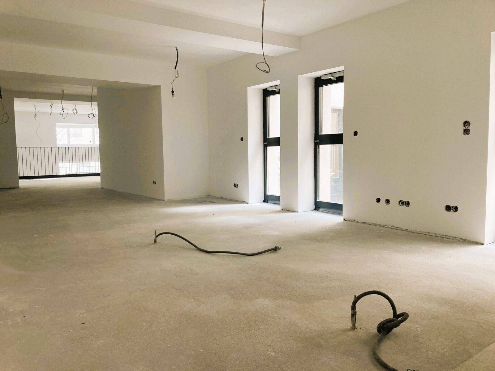 Geräumiger, unfertiger Innenraum mit weißen Wänden, Betonboden und großen Fenstertüren, bereit für den Ausbau.