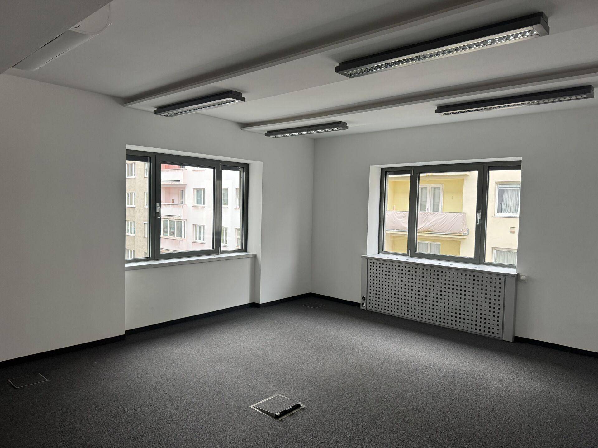 Heller Büroraum mit Teppichboden, großen Fenstern und Blick auf andere Gebäude.