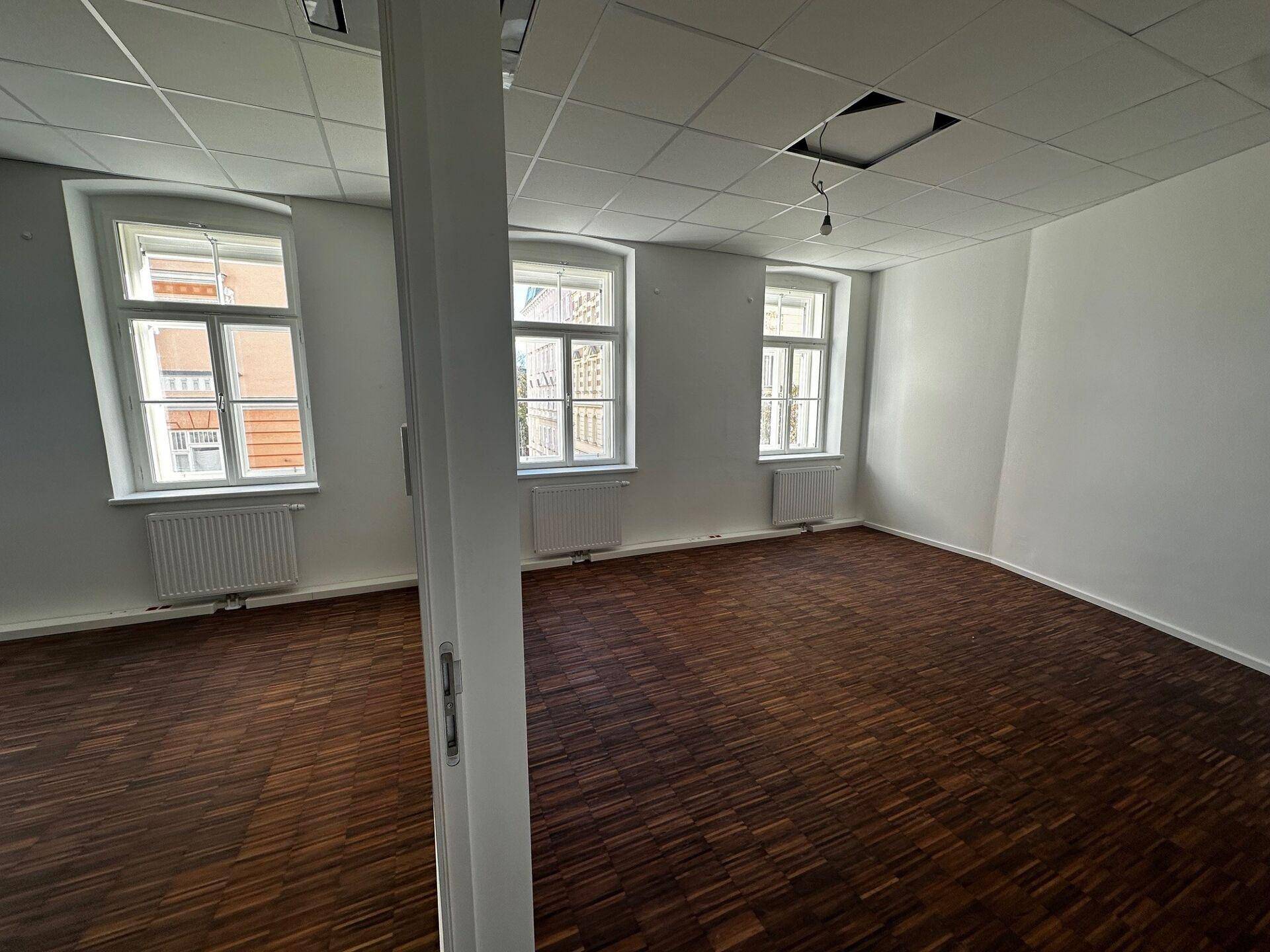 Helles, leeres Büro mit Holzboden, weißen Wänden und drei Fenstern mit Blick nach draußen.