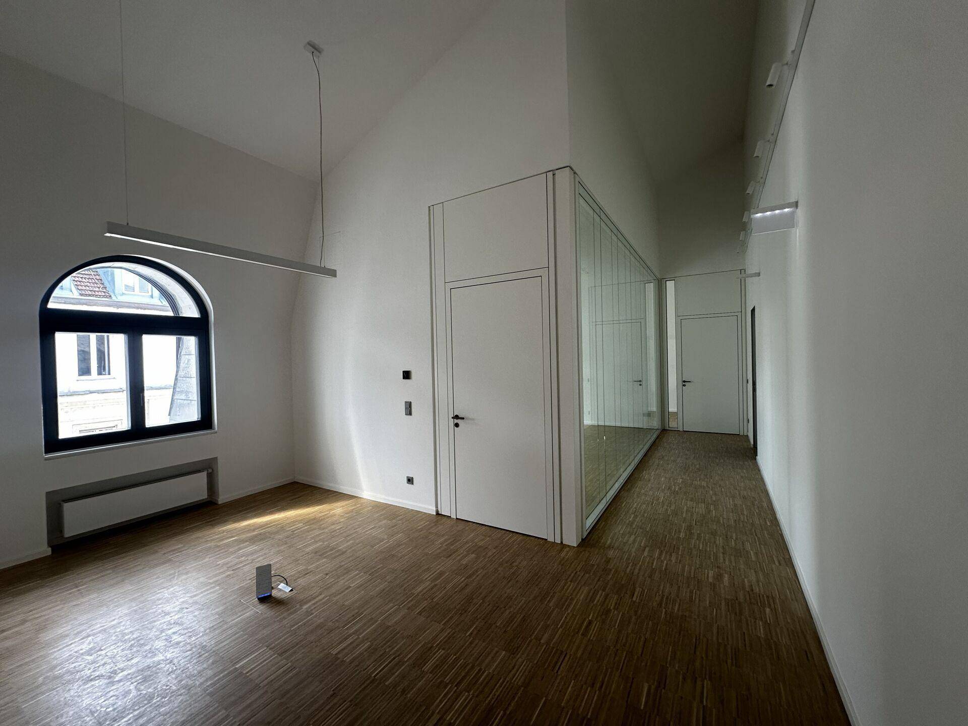 Heller Büroraum mit Holzboden, einem Rundbogenfenster und einer modernen Glaswand als Raumteiler.