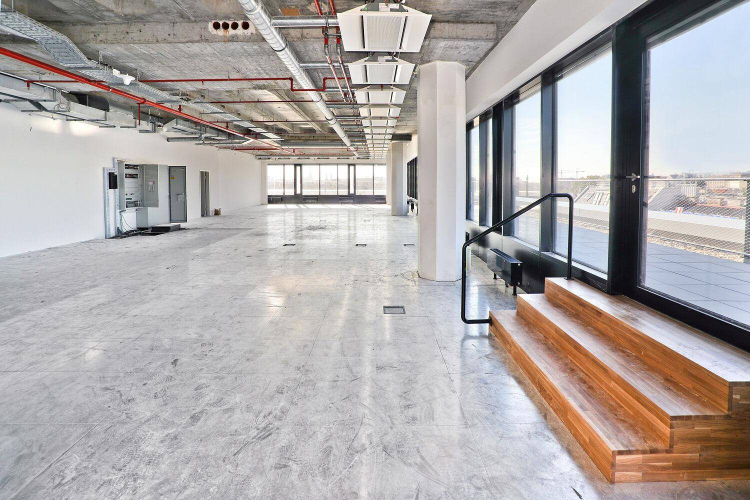 Offener Bürobereich mit Betonboden, großen Fenstern und einer modernen Holztreppe, die zu einer Außenterrasse führt und den Raum verbindet.