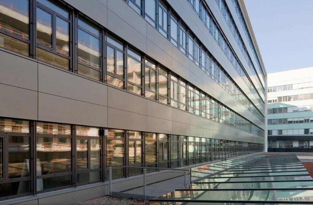 Lange Fassade eines modernen Bürogebäudes mit vielen Fenstern und einem gläsernen Dach über einem Außenbereich.