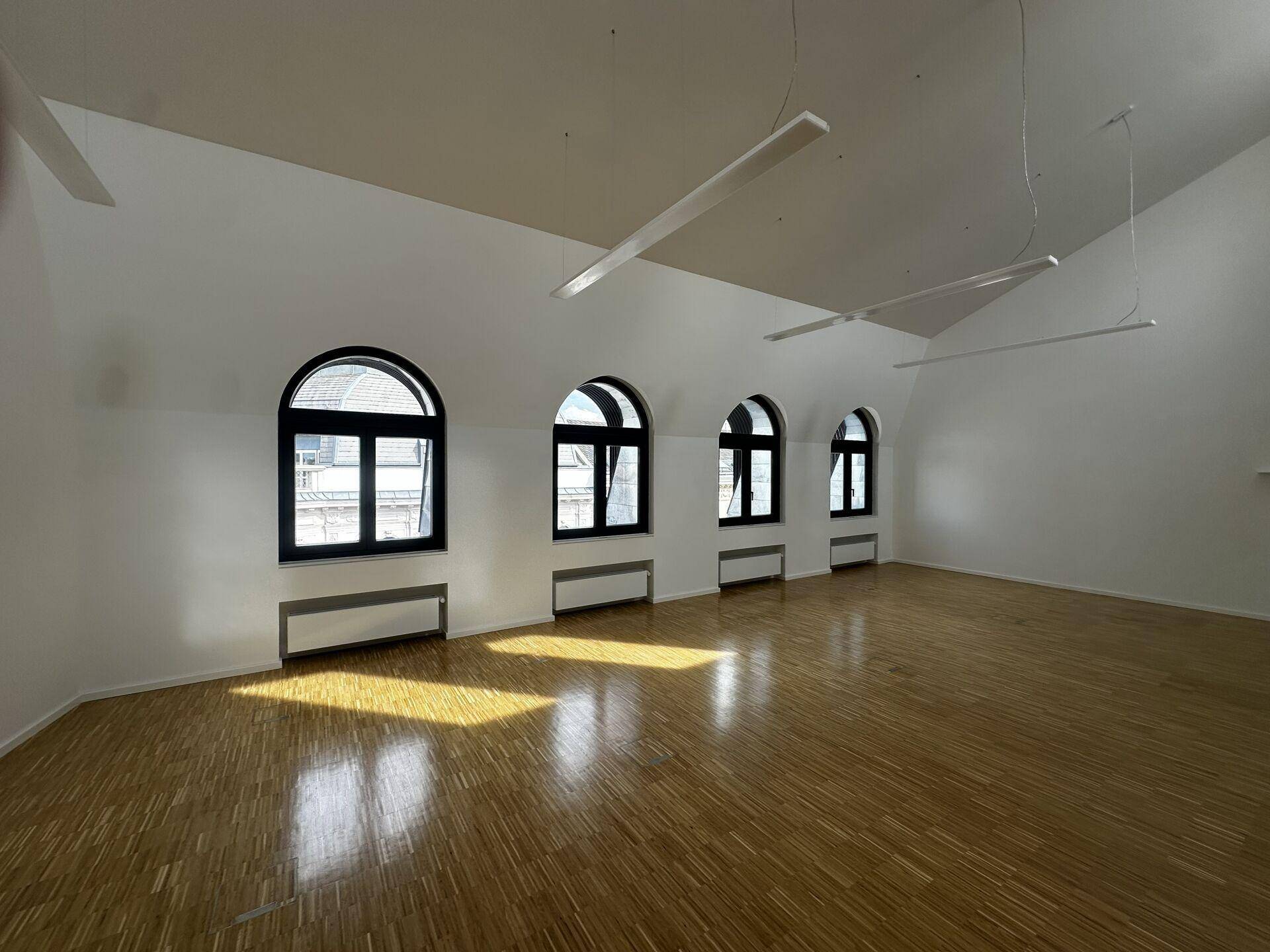 Heller Büroraum mit Holzboden, schrägen Decken und vier eleganten Rundbogenfenstern, die viel Licht hereinlassen.