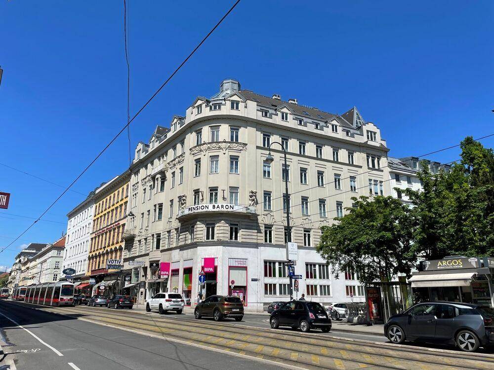 Historisches Gebäude mit Geschäften im Erdgeschoss und Straßenbahnlinien in einer belebten Stadtansicht.