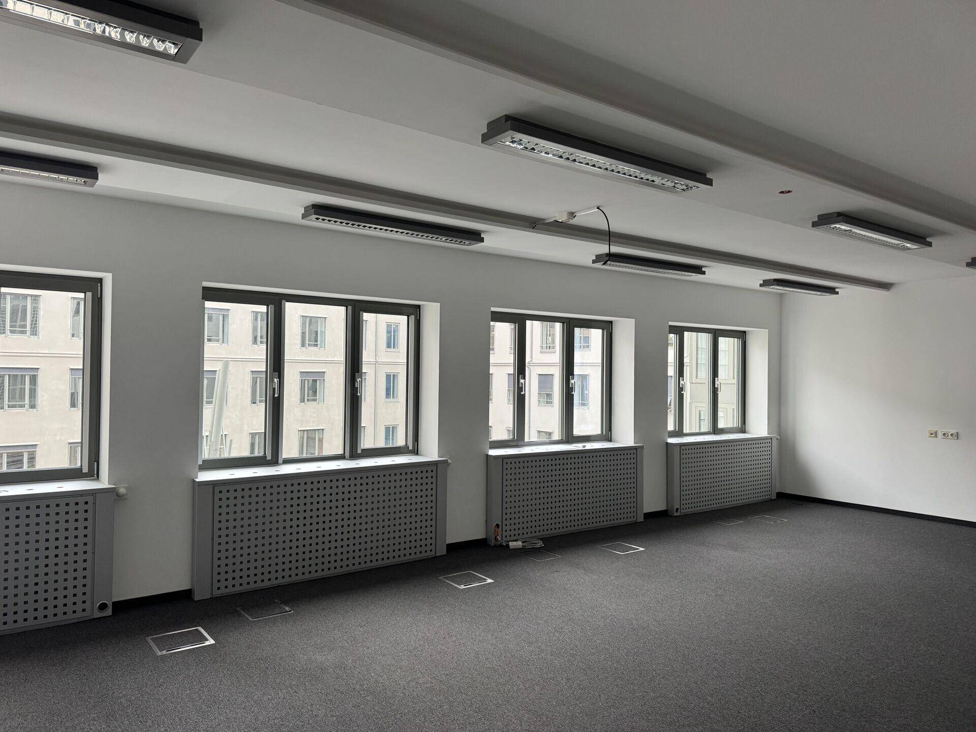 Langes Büro mit mehreren großen Fenstern, die viel Tageslicht hereinlassen, Teppichboden und moderner Beleuchtung.