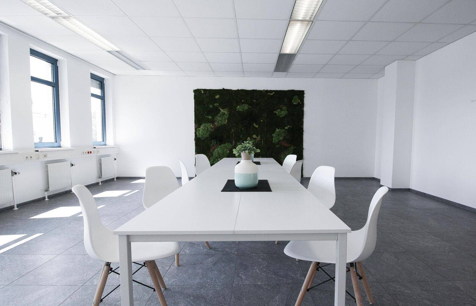 Heller Konferenzraum mit großem weißen Tisch, modernen Stühlen und einer dekorativen grünen Mooswand.