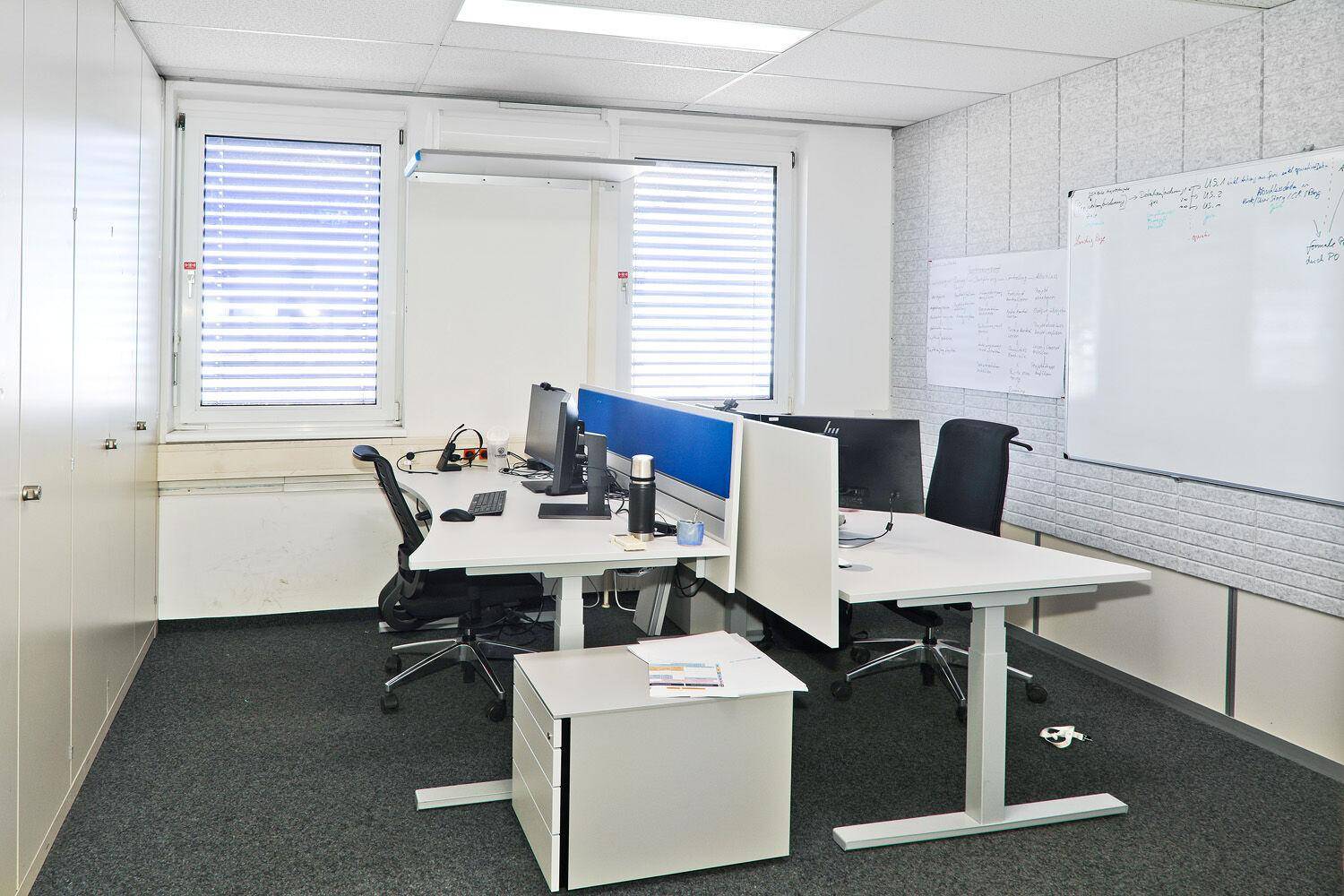 Modernes Büro mit zwei Arbeitsplätzen, weißen Schränken und einer großen Whiteboard-Wand.