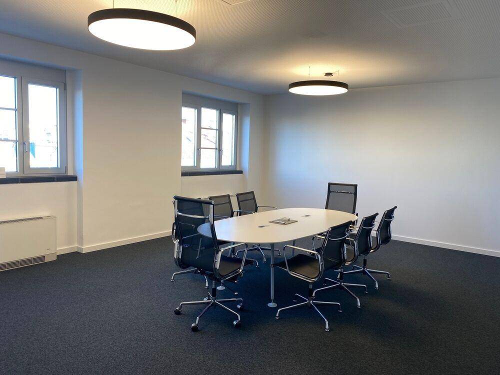 Moderner Konferenzraum mit ovalem Tisch, Bürostühlen und dunklem Teppichboden.