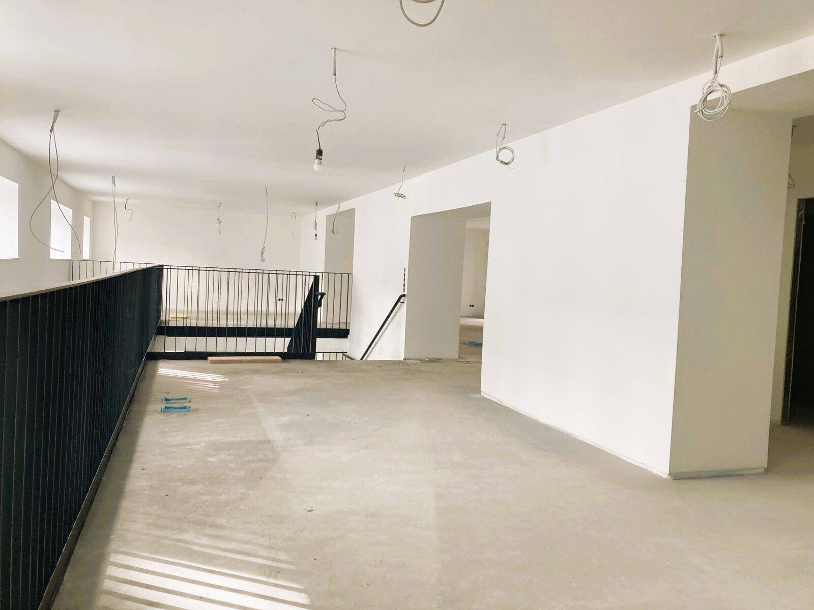 Großer, heller Innenraum im Rohbau mit schwarzem Geländer einer Galerie und Betonboden, bereit für den Ausbau.