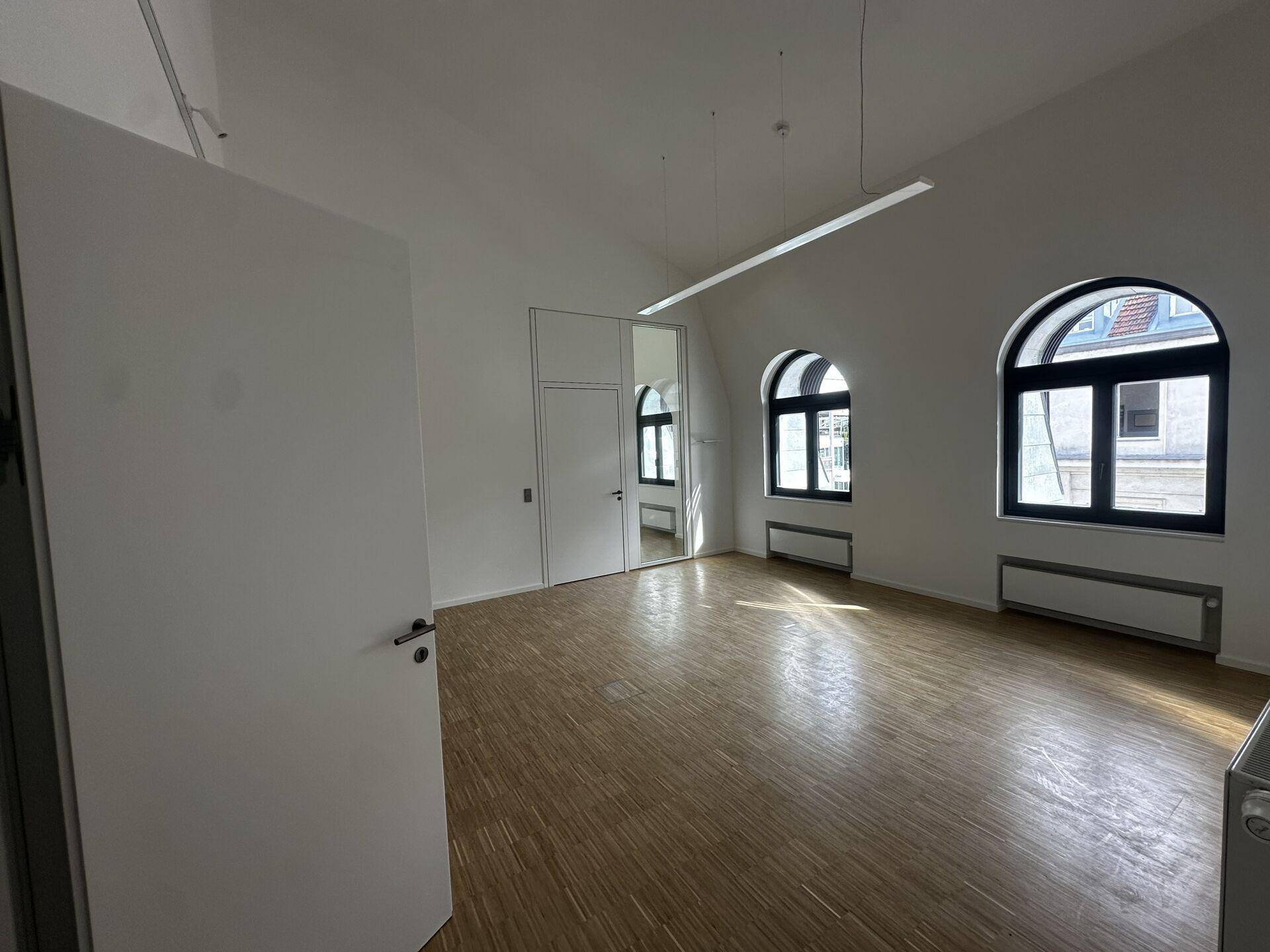 Heller Büroraum mit Holzboden, zwei Rundbogenfenstern und einer modernen Spiegelfläche an der Wand.