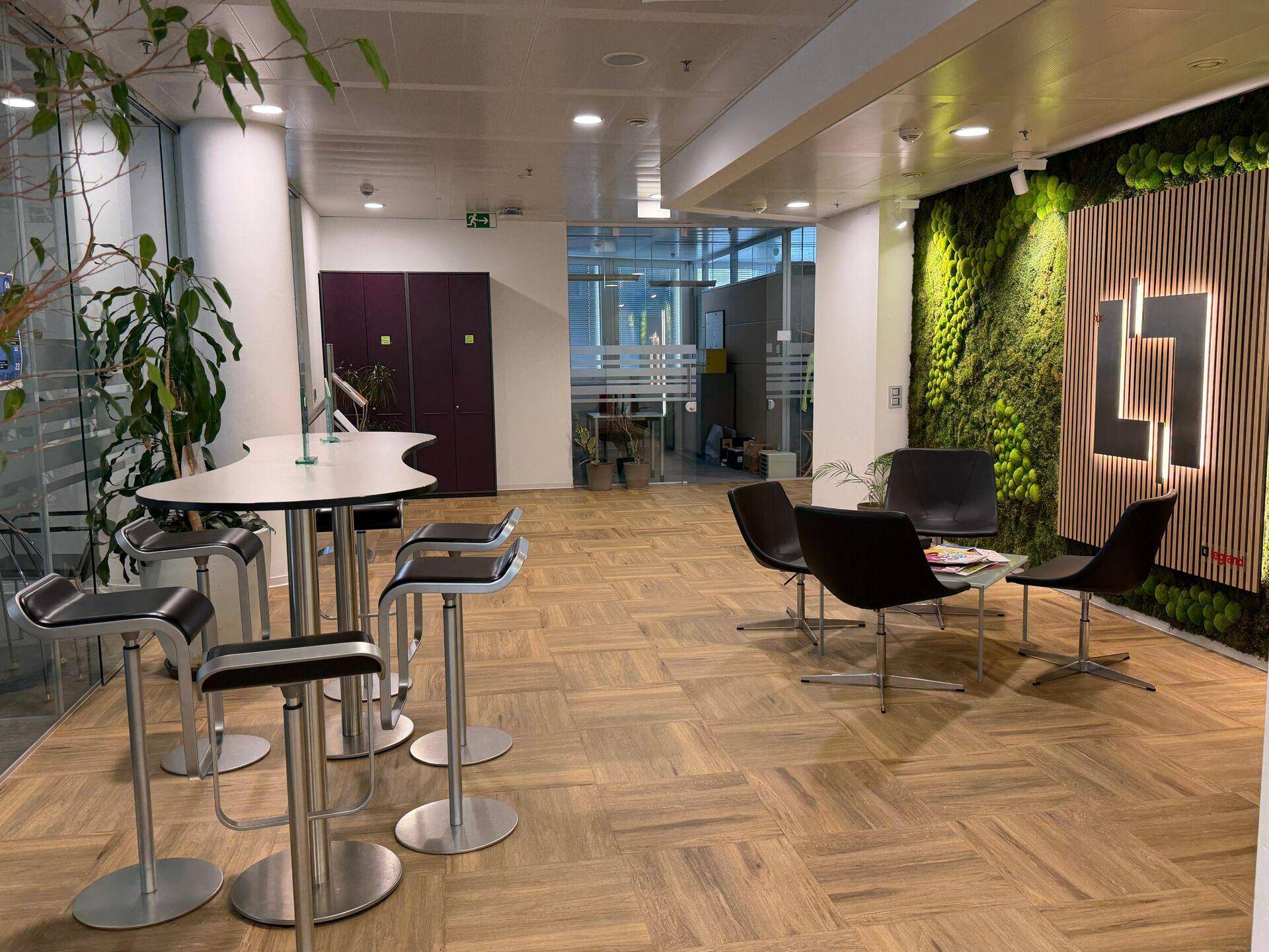 Moderner Gemeinschaftsbereich mit Holzboden, Stehtischen und einer Lounge-Ecke, akzentuiert durch eine Mooswand.