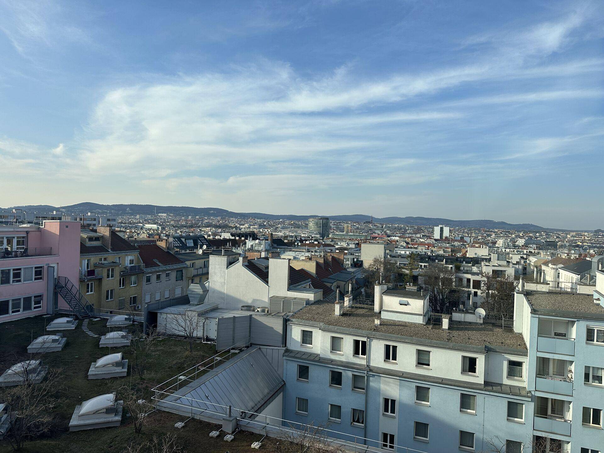Weitläufiger Blick über eine Stadtlandschaft mit Wohngebäuden, Grünflächen und Bergen am Horizont.