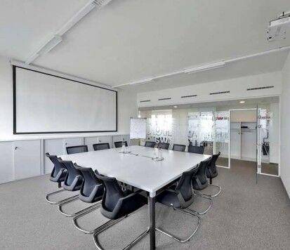 Heller Konferenzraum mit großem weißen Tisch, schwarzen Stühlen, Projektionsfläche und Glaswänden.