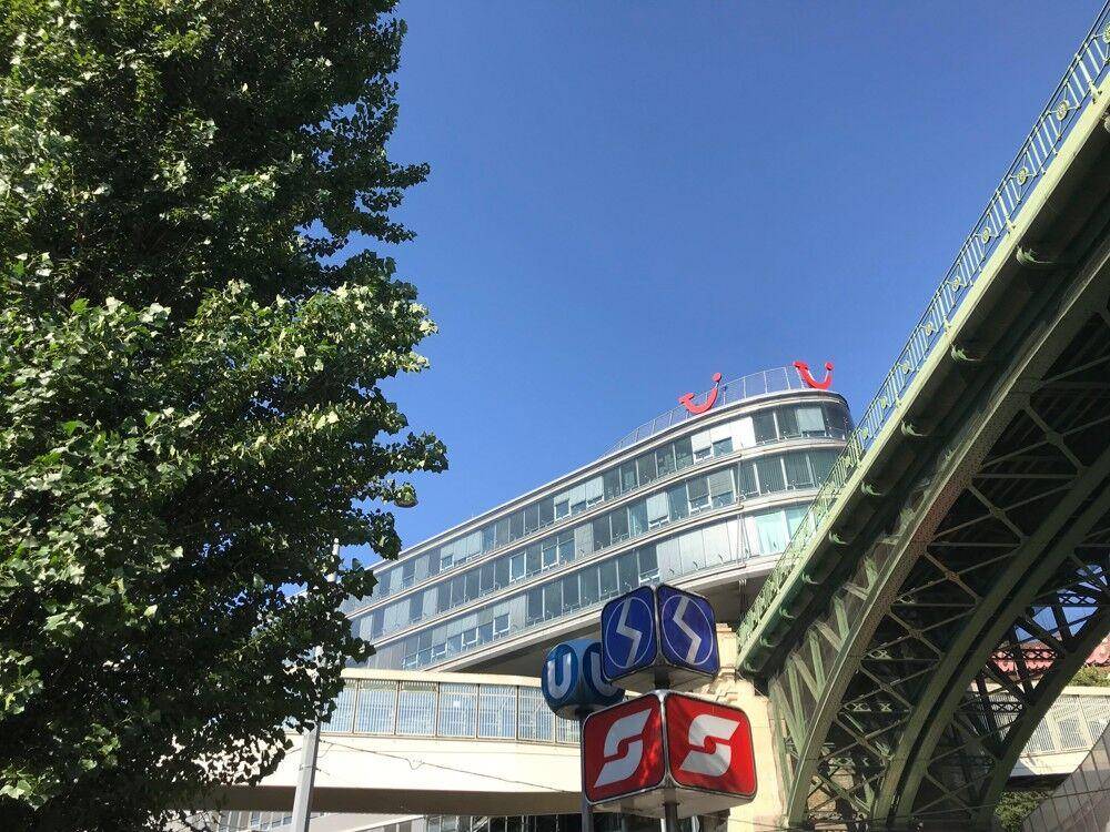 Modernes Gebäude mit U-Bahn-Schildern und grüner Brücke, umgeben von Bäumen unter klarem Himmel.