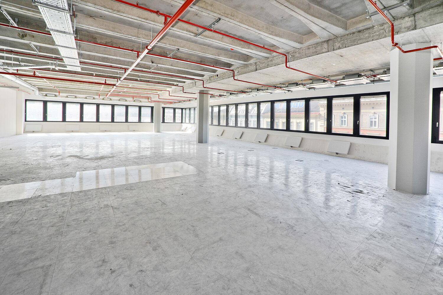 Weitläufiger Bürobereich im Rohzustand mit Betonboden, hohen Decken und großen Fenstern, die eine helle und offene Arbeitsumgebung schaffen.