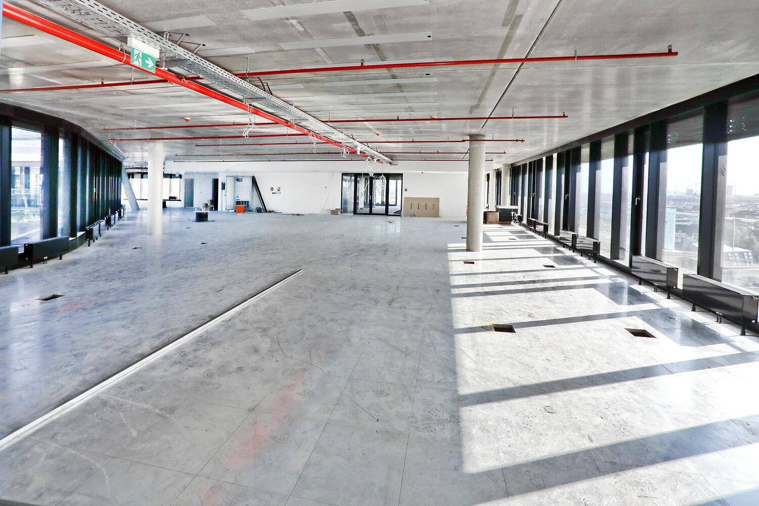 Großer, offener Büroraum im Rohbau mit Betonboden, sichtbaren Installationen und großen Fenstern mit Stadtblick.
