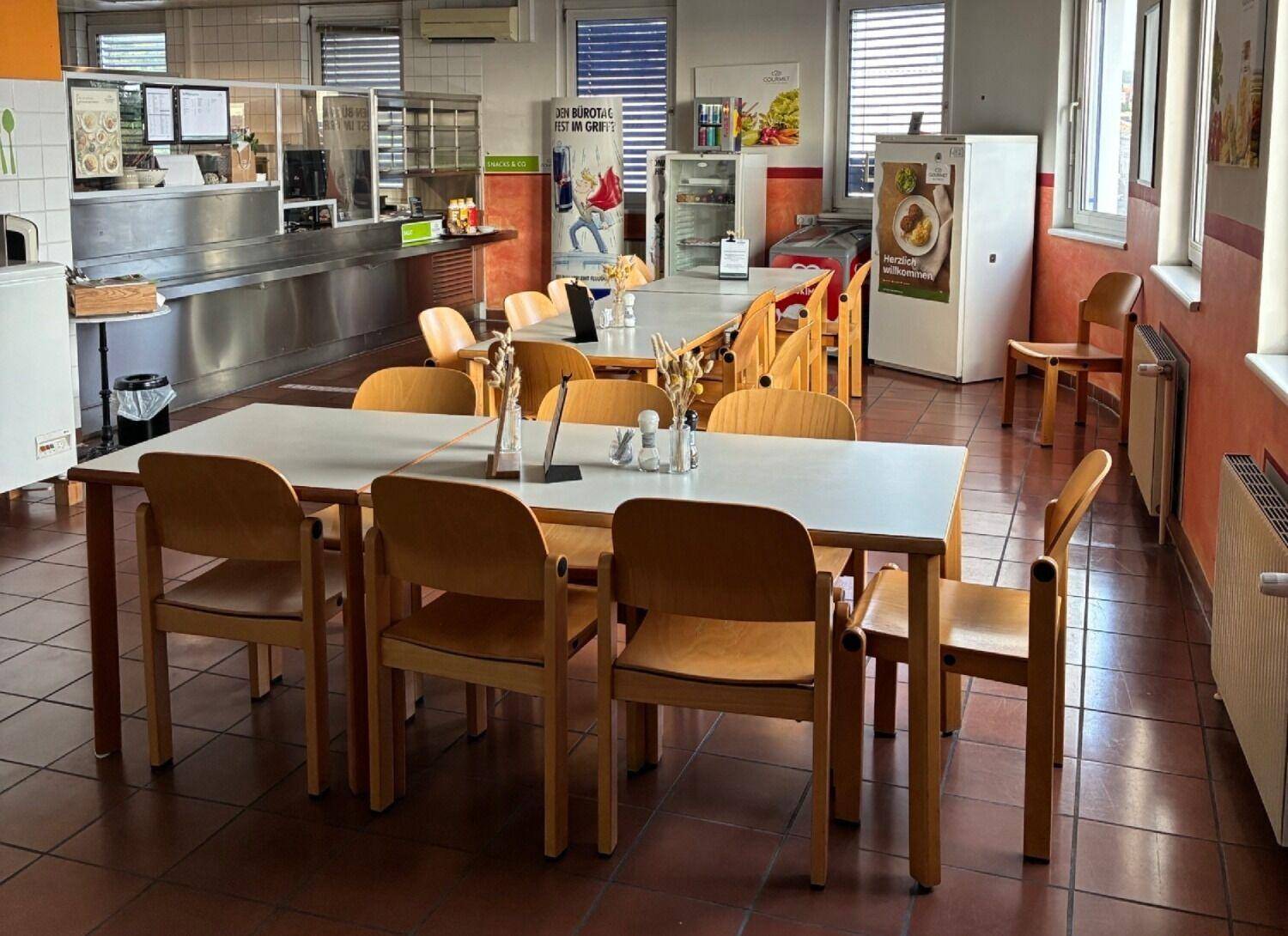 Kantinenbereich mit Holztischen und Stühlen, Fliesenboden und einer Essensausgabe im Hintergrund.