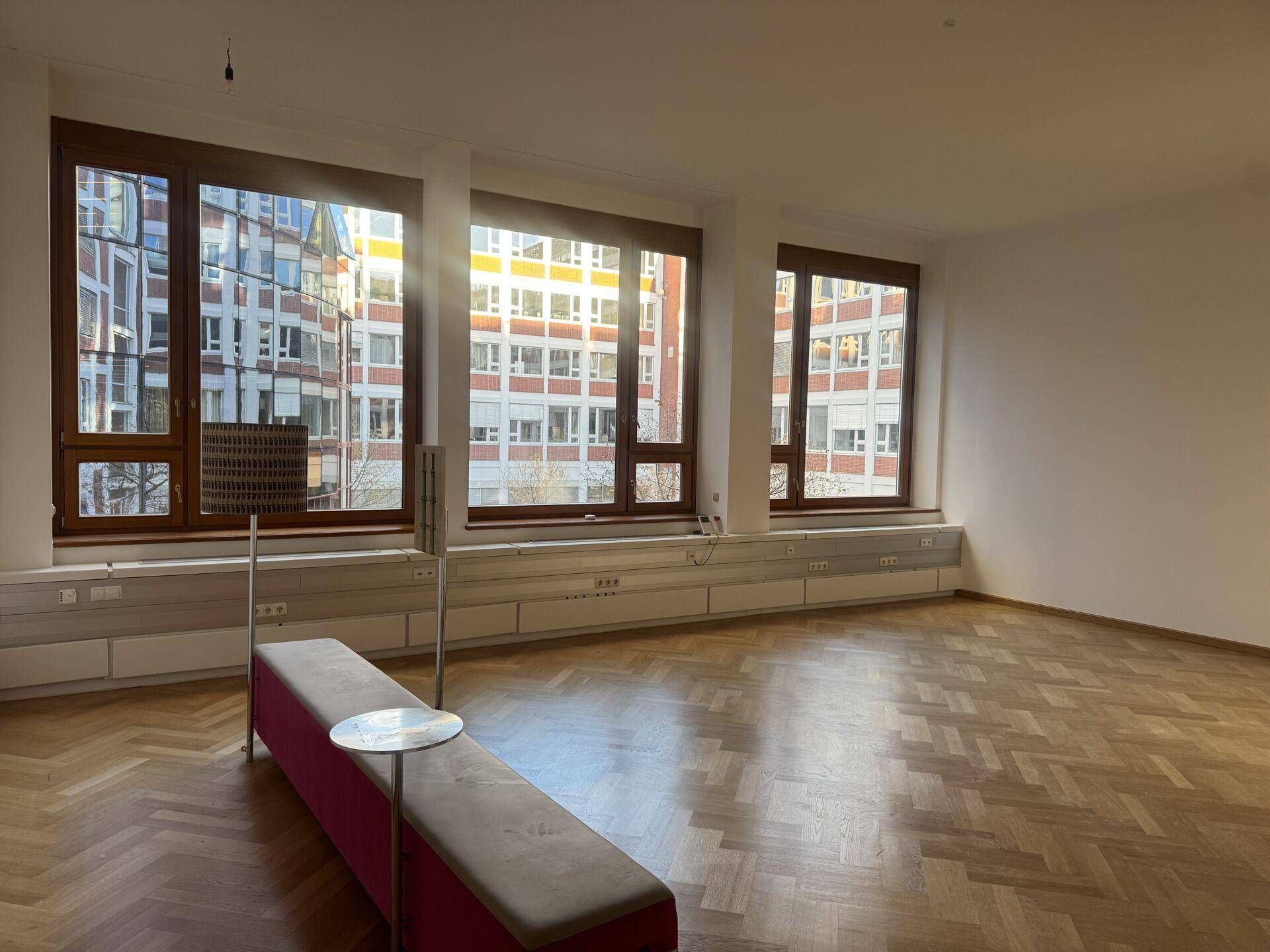 Geräumiger, heller Büroraum mit großen Fenstern, Fischgrätparkett und Blick auf umliegende Gebäude.