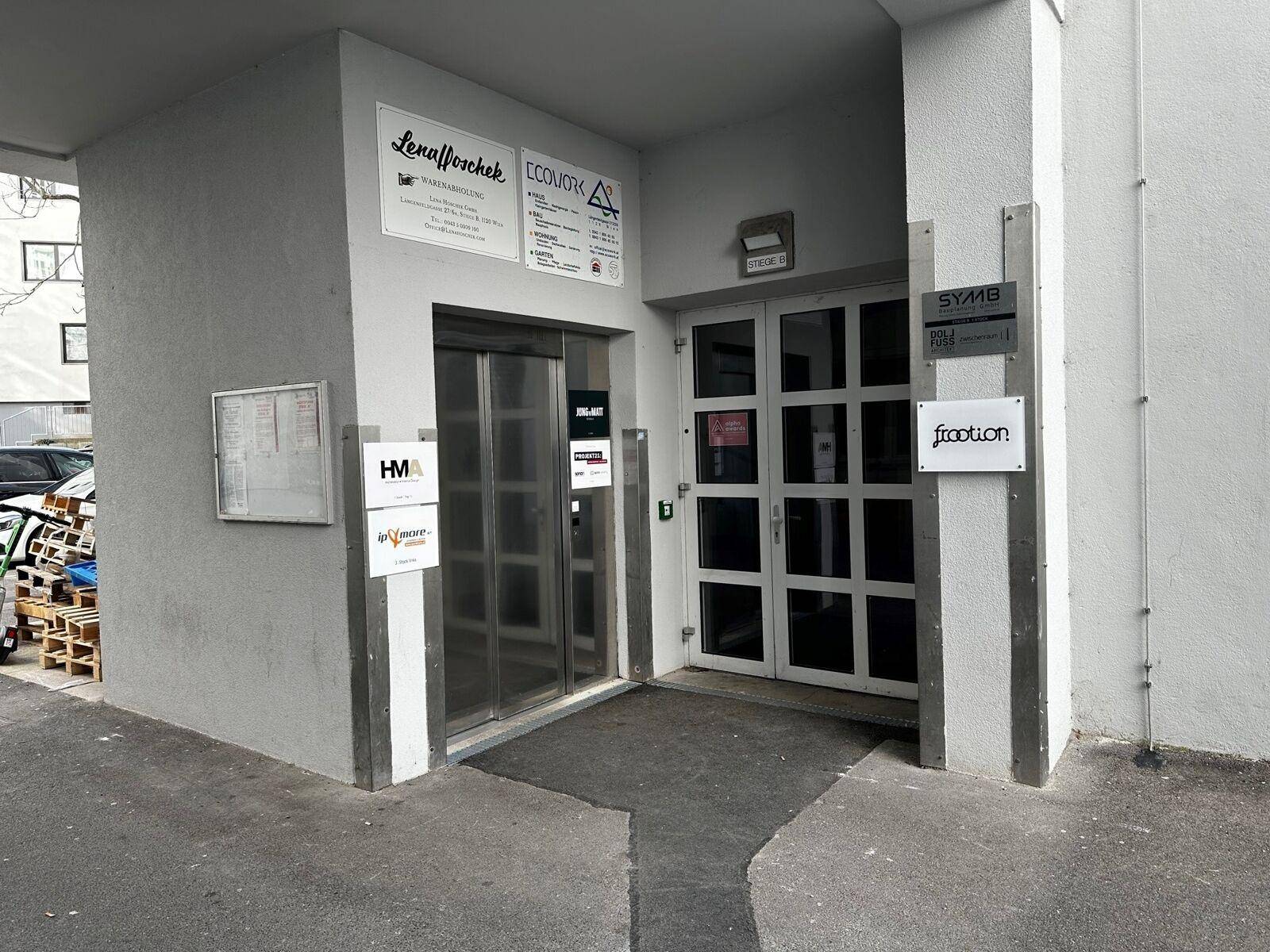 Eingangsbereich mit Aufzug und Glastür, umgeben von hellgrauen Wänden und Geschäftsschildern.