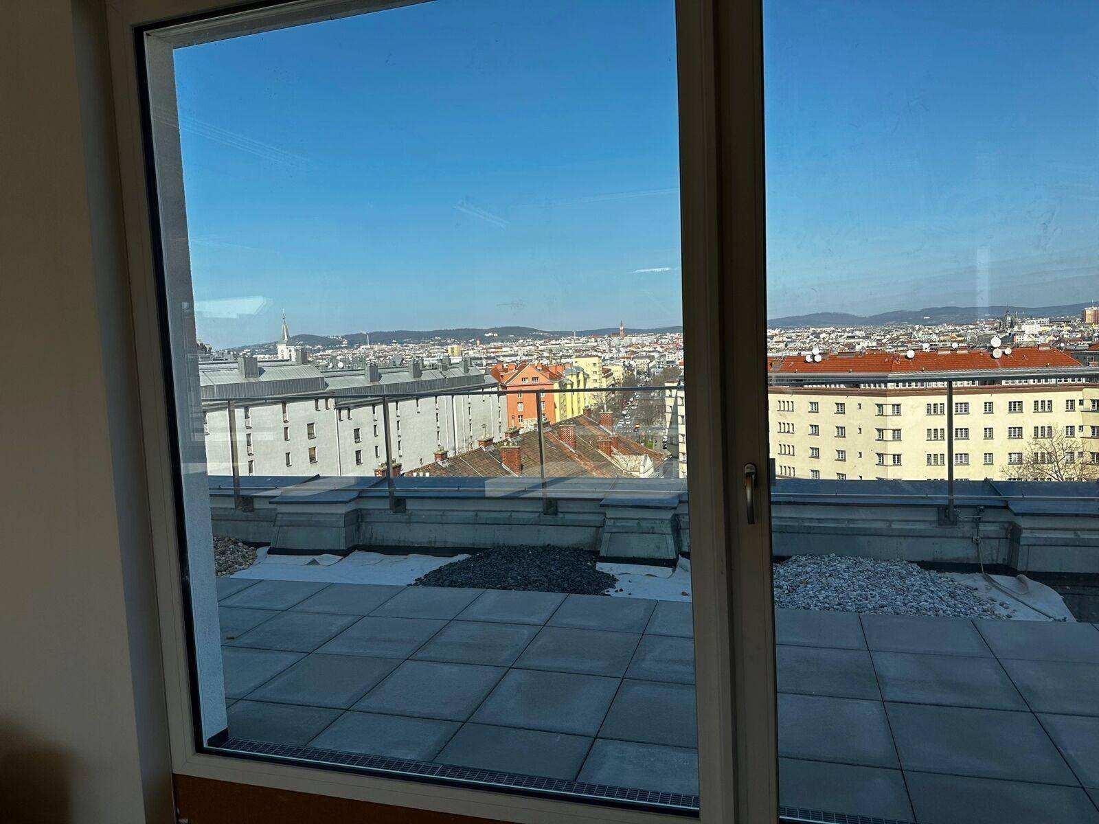 Blick aus einem Fenster auf eine Dachterrasse und die weite Stadtlandschaft unter blauem Himmel.