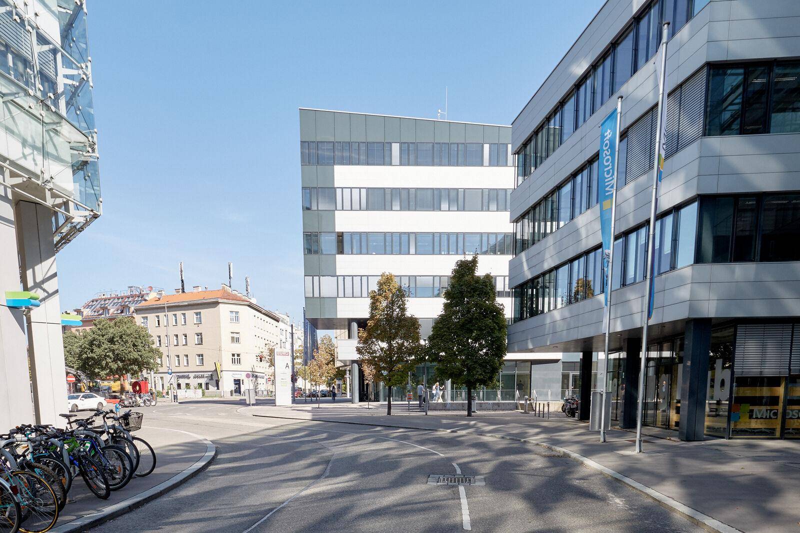 Eckansicht eines modernen Bürogebäudes an einer Straße mit Fahrradständern und weiteren Gebäuden im Hintergrund.
