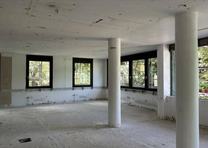 Unfertiger Innenraum mit Betonboden, tragenden Säulen und großen Fenstern, die viel natürliches Licht hereinlassen.