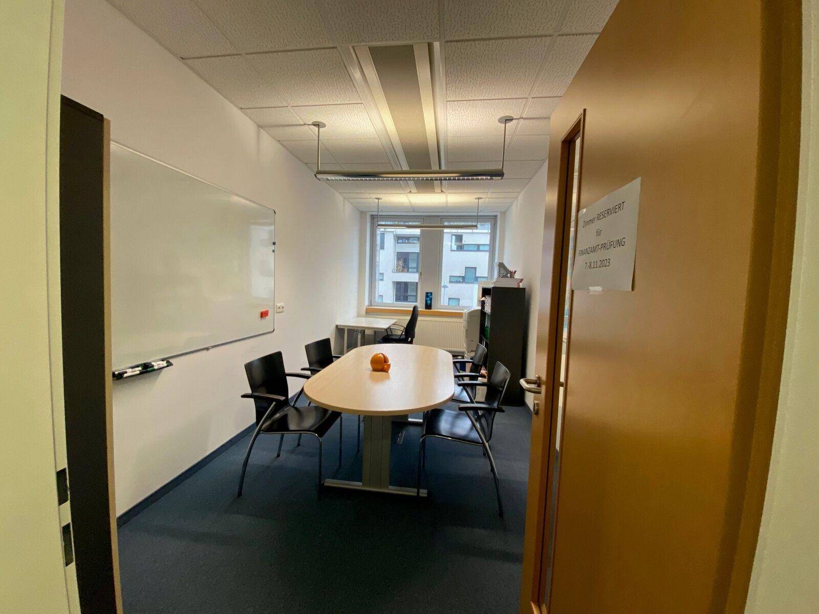 Kleiner Besprechungsraum mit ovalem Tisch, Stühlen, Whiteboard und Fensterblick in eine städtische Umgebung.