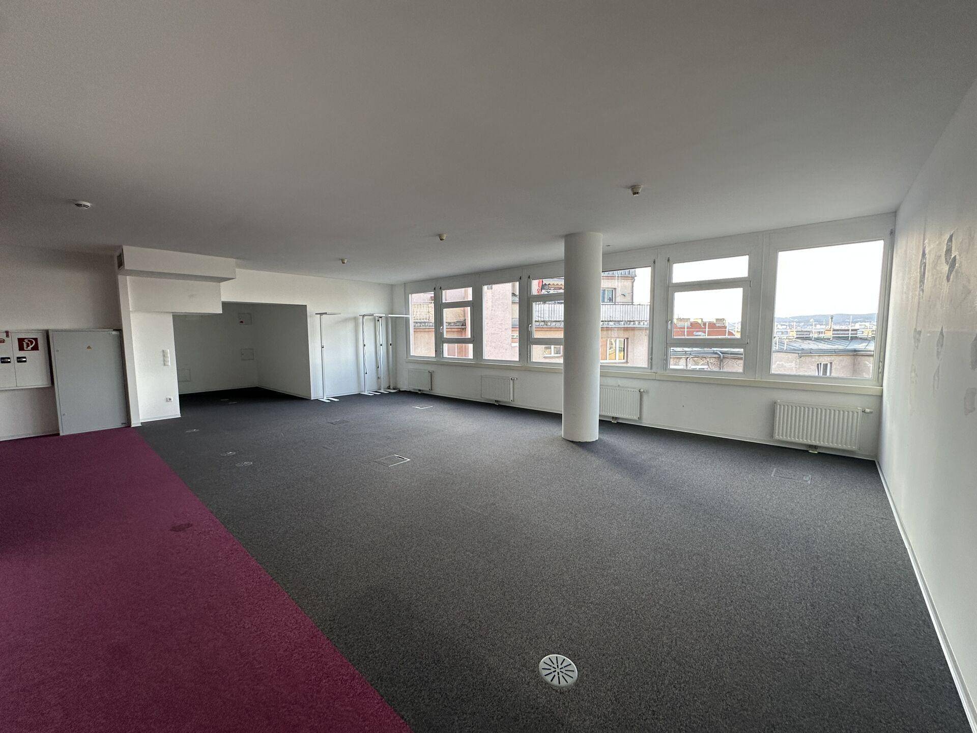 Großer, offener Büroraum mit Teppichboden, vielen Fenstern und einer Säule in der Mitte.