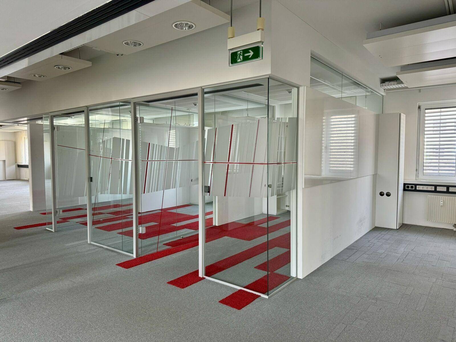 Modernes Büro mit Glasabtrennungen, roten Akzenten auf dem Teppich und viel Tageslicht.