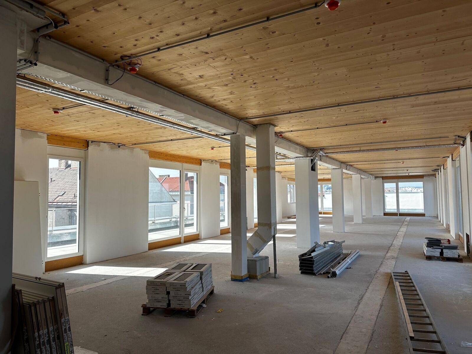 Großer, offener Raum im Rohbau mit Holzbalkendecke, Betonboden und vielen Fenstern, die viel Licht hereinlassen.
