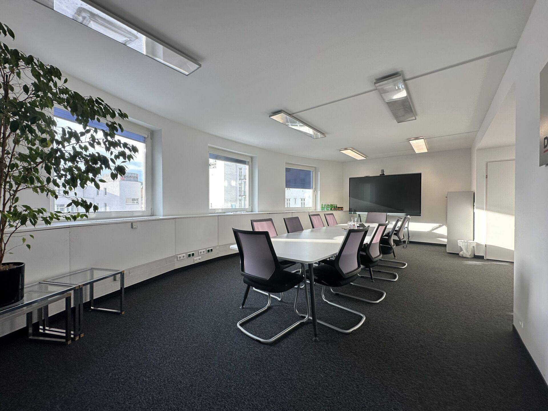 Heller Konferenzraum mit großem Tisch, modernen Stühlen und einem großen Bildschirm an der Wand.