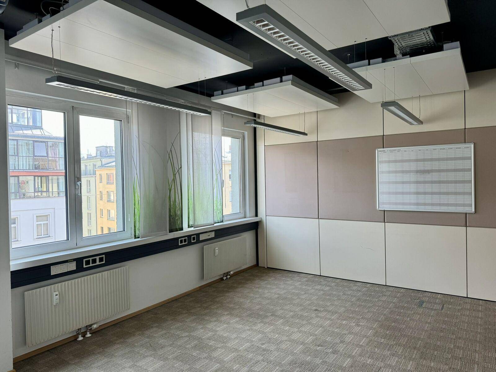 Leeres Büro mit großen Fenstern, Heizkörpern und schallabsorbierenden Deckenpaneelen.