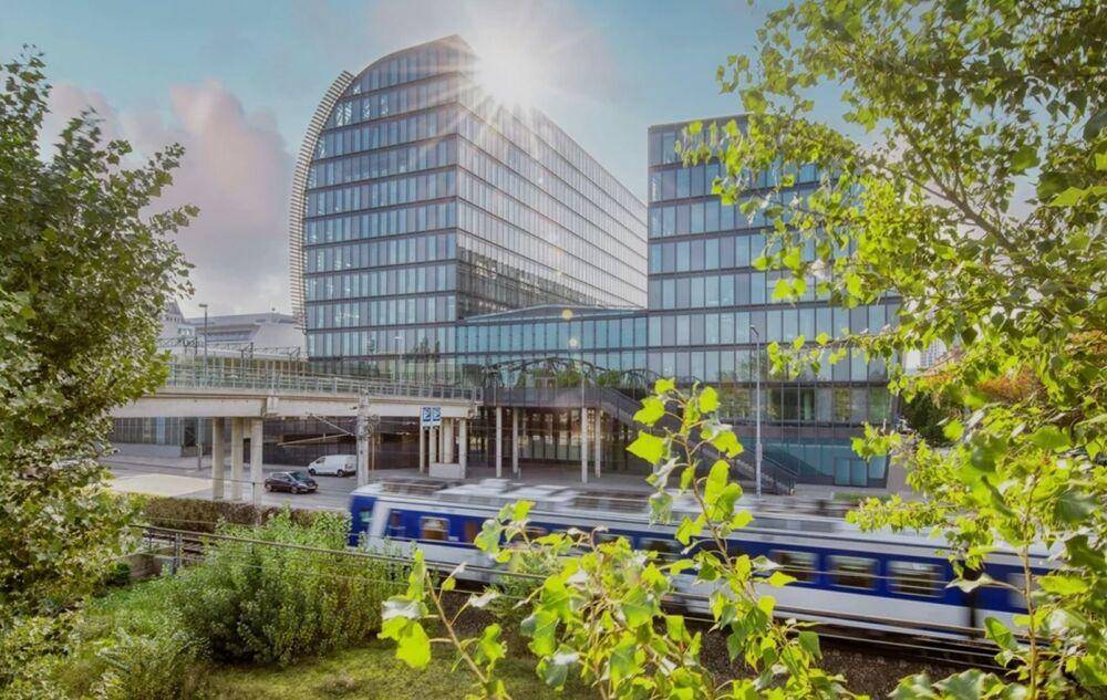 Modernes Bürogebäude mit reflektierender Glasfassade, umgeben von Bäumen und einer vorbeifahrenden Bahn.